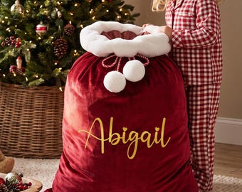 May include: A large, deep red velvet Christmas gift sack featuring a white faux fur trim at the top, two white pom-poms on red and white twisted cords, and the name Abigail printed in gold script lettering across the front of the fabric.