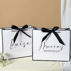 May include: Two white gift bags with black ribbon bows and black trim. The bag on the left says "Louise" and the bag on the right says "Murray Bridesmaid".