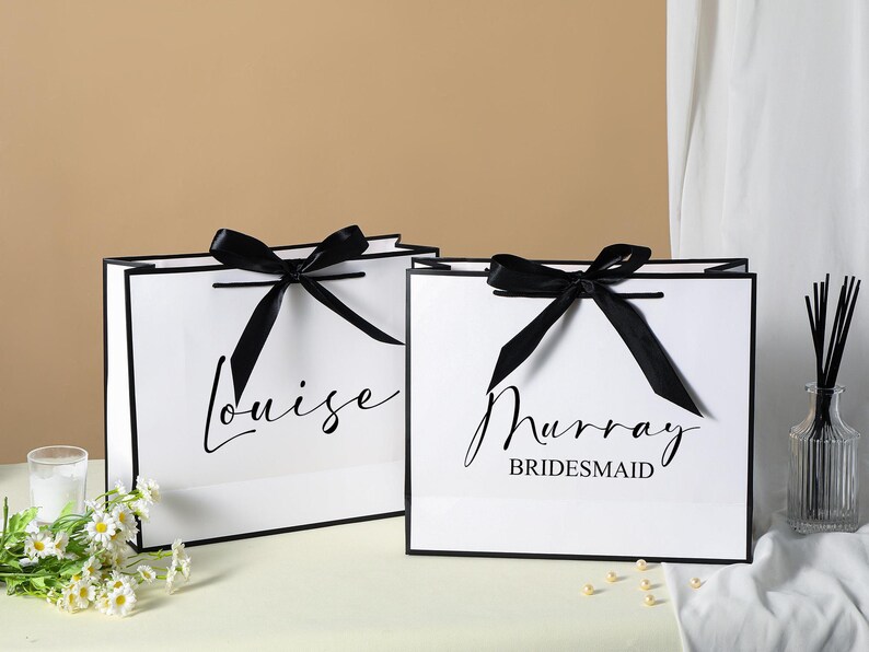 May include: Two white gift bags with black ribbon bows and black trim. The bag on the left says "Louise" and the bag on the right says "Murray Bridesmaid".