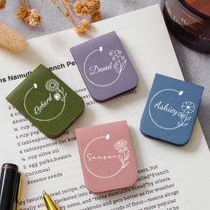 May include: Four personalized leather bookmarks in green, purple, pink, and blue, each with a name and floral design. The bookmarks are on a book page with text and a pen.