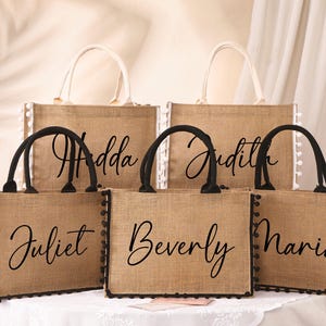 Op de afbeelding: Vier bruine jute tassen met zwarte pomponrand en zwarte kalligrafie. De tassen zijn gelabeld met "Hadda", "Judith", "Juliet" en "Maria".
