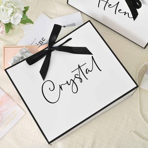 May include: White gift bags with black trim and black ribbon bows. The word "Crystal" is written in cursive on one bag, and "Helen" on another. The bags are surrounded by small gift boxes, flowers, and a pearl necklace.