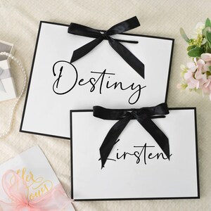 May include: Two white gift bags with black trim and black bows. The word "Destiny" is written on the larger bag, and "Kirsten" on the smaller one. A small gift box with a silver bow and a pearl necklace are also visible.