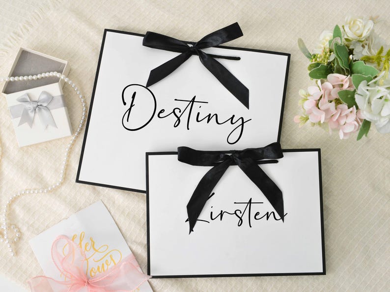 May include: Two white gift bags with black trim and black bows. The word "Destiny" is written on the larger bag, and "Kirsten" on the smaller one. A small gift box with a silver bow and a pearl necklace are also visible.