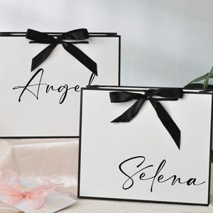 May include: Two white gift bags with black trim and black bows. The bags have the names "Angel" and "Selena" written in elegant script. The bags are on a white surface with a pink ribbon and flowers.