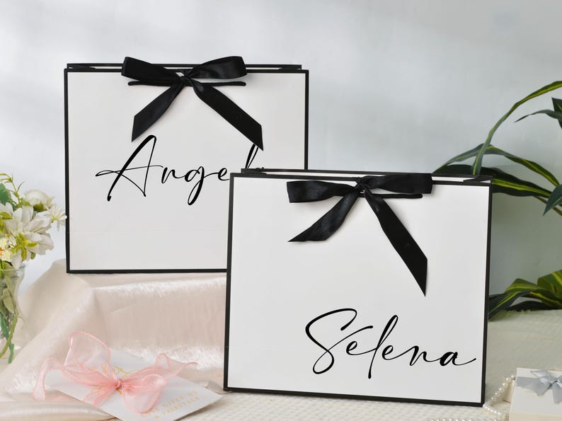 May include: Two white gift bags with black trim and black bows. The bags have the names "Angel" and "Selena" written in elegant script. The bags are on a white surface with a pink ribbon and flowers.