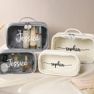 Op de afbeelding: Vier transparante cosmetica tassen met zilveren ritsen en handvatten. Twee tassen zijn grijs, met de tekst "Jessica", en twee zijn wit, met de tekst "Sophia".