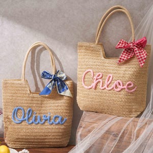 May include: Two woven straw tote bags with dual handles. The left bag features the name Olivia in blue script lettering and a blue patterned ribbon. The right bag features the name Chloe in pink script lettering and a red and white checkered ribbon.