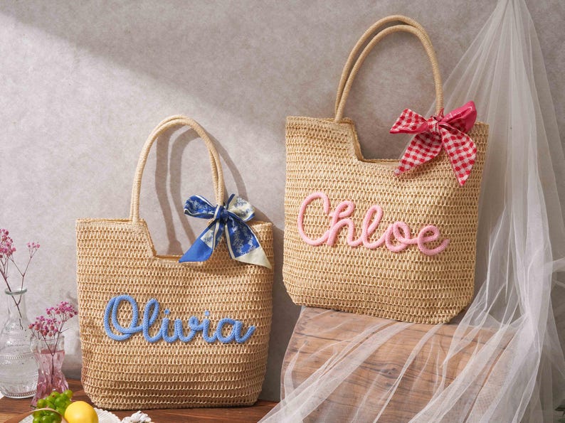 May include: Two woven straw tote bags with dual handles. The left bag features the name Olivia in blue script lettering and a blue patterned ribbon. The right bag features the name Chloe in pink script lettering and a red and white checkered ribbon.