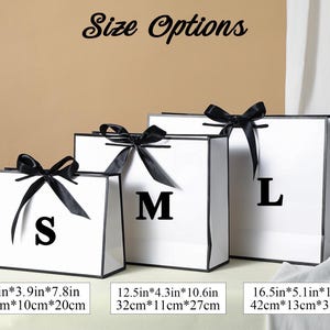 May include: Three white gift bags with black ribbon bows and black trim. The bags are labelled with the letters S, M, and L. The bags are 28 cm by 10 cm by 20 cm, 32 cm by 11 cm by 27 cm, and 42 cm by 13 cm by 31 cm.