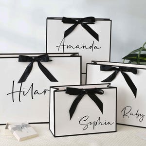 May include: White gift bags with black trim and bows. Each bag has a name written in cursive: Amanda, Hilar, Sophia, and Ruby. The bags are arranged on a white surface, with a small gift box and flowers in the background.