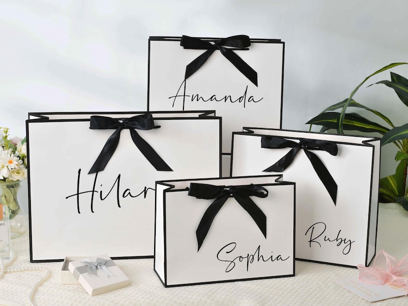 May include: White gift bags with black trim and bows. Each bag has a name written in cursive: Amanda, Hilar, Sophia, and Ruby. The bags are arranged on a white surface, with a small gift box and flowers in the background.