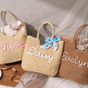 May include: Three woven straw tote bags with two top handles each. The bags feature personalized names in raised script lettering: Chloe in pink, Daisy in white, and Evelyn in white. Each bag is accented with a decorative fabric bow tied to one handle.