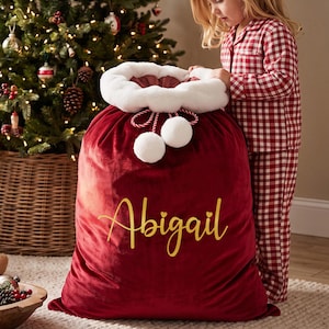 May include: A large red velvet Santa sack featuring a white faux fur trim at the top, two white pom-poms on red and white twisted cords, and the name Abigail printed in gold cursive script on the front of the fabric.