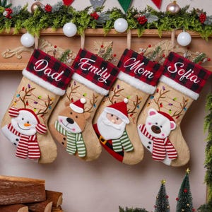 Könnte beinhalten: Vier Weihnachtsstrümpfe aus Jute mit rot-schwarz karierten Bündchen, personalisiert mit den Namen Dad, Emily, Mom und Sofia. Die Strümpfe sind mit gestickten Rentiergeweihen sowie Plüschapplikationen eines Schneemanns, Rentiers, Weihnachtsmanns und Eisbären verziert.
