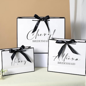 May include: Three white gift bags with black ribbon bows and black trim. The bags are labelled with the names "Cheryl Bridesmaid", "Athena Bridesmaid", and "Hilary".