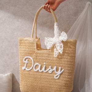 May include: A beige woven straw tote bag featuring the name Daisy embroidered in white script lettering on the front. The bag includes two matching woven straw handles and a decorative white lace ribbon tied to one of the handles near the top.