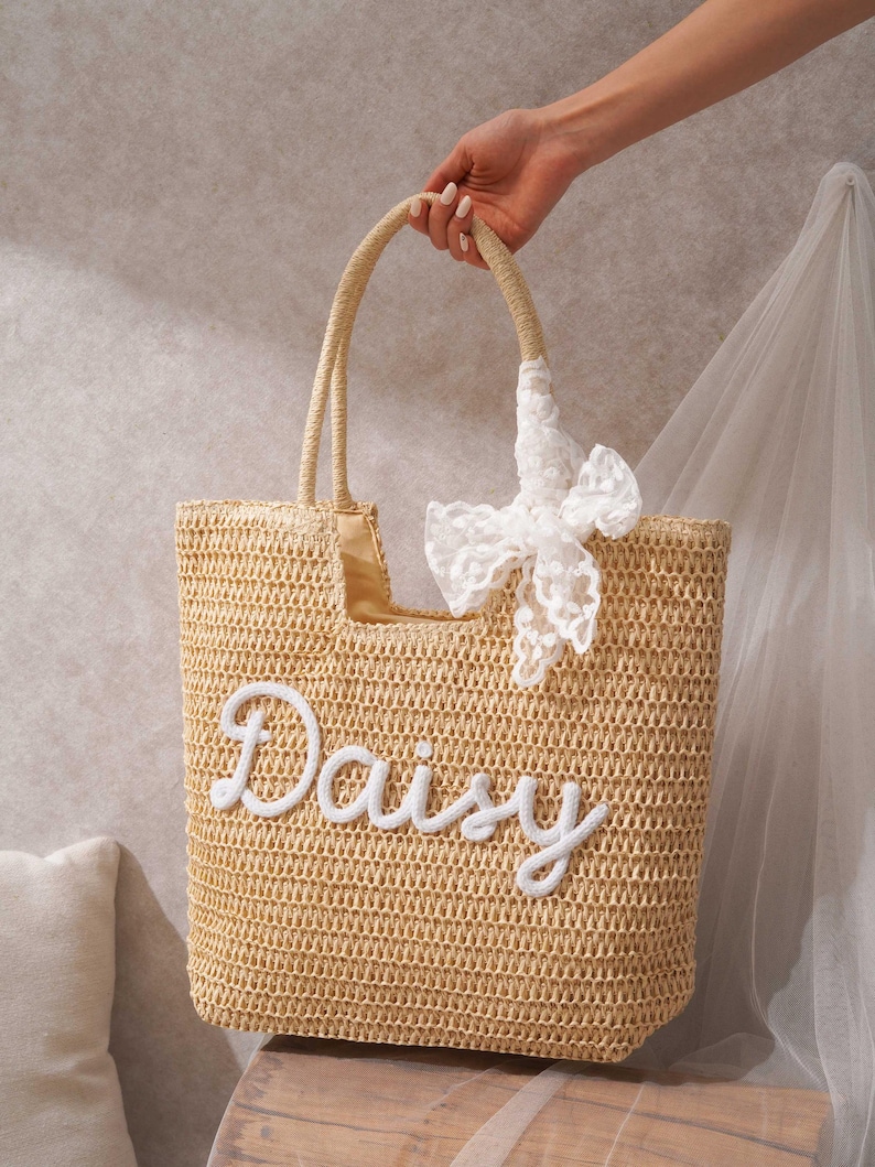 May include: A beige woven straw tote bag featuring the name Daisy embroidered in white script lettering on the front. The bag includes two matching woven straw handles and a decorative white lace ribbon tied to one of the handles near the top.