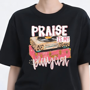 Può includere: T-shirt nero con grafica di un giradischi vintage sopra una valigia rosa. Il testo "PRAISE IS MY PLAYLIST" è visualizzato in un carattere giocoso, con una combinazione di colori rosa e bianco. Il design ha un'estetica retrò a tema musicale.