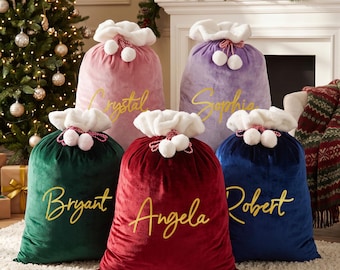 May include: Five velvet Christmas gift sacks in pink, purple, dark green, red, and navy blue. Each sack features a white plush cuff with two white pom-poms on a red and white cord. The names Crystal, Sophia, Bryant, Angela, and Robert are in gold.