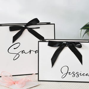 May include: Two white gift bags with black trim and black bows. The larger bag has the name "Sara" in cursive, and the smaller bag has "Jessica". A pink ribbon and a white card are in the foreground.