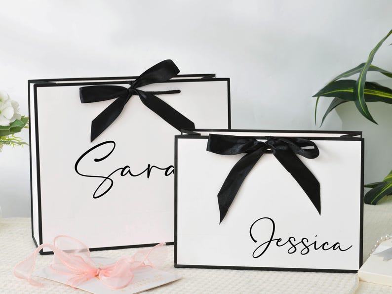 May include: Two white gift bags with black trim and black bows. The larger bag has the name "Sara" in cursive, and the smaller bag has "Jessica". A pink ribbon and a white card are in the foreground.
