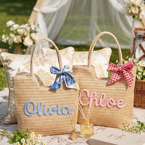 May include: Two woven straw tote bags with top handles. One bag features the name Olivia in blue script with a blue patterned ribbon. The other bag features the name Chloe in pink script with a red and white checkered ribbon tied to the handle.