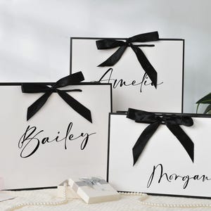 May include: Three white gift bags with black trim and black satin bows. Each bag has a name written in cursive: Amelia, Bailey, and Morgan. A small gift box and a string of pearls are in front of the bags.