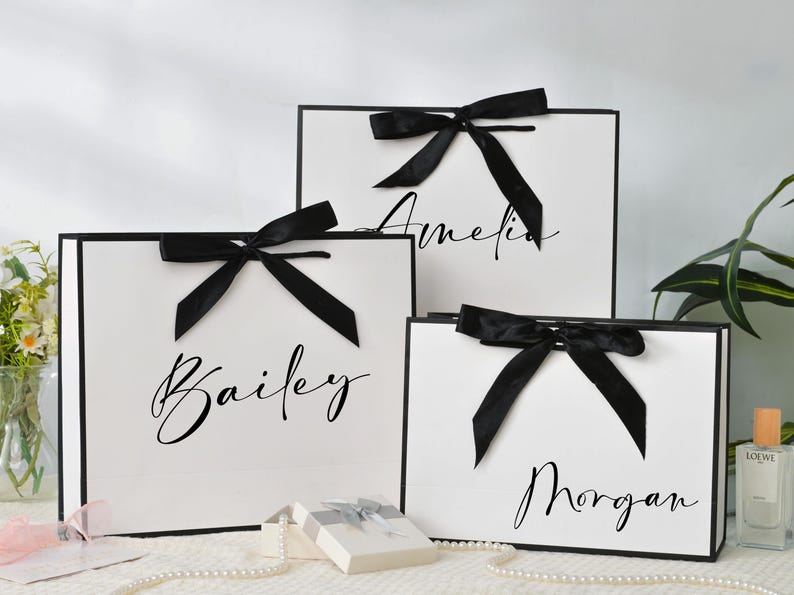 May include: Three white gift bags with black trim and black satin bows. Each bag has a name written in cursive: Amelia, Bailey, and Morgan. A small gift box and a string of pearls are in front of the bags.