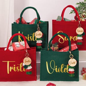 Könnte beinhalten: Vier Tragetaschen in Rot und Grün, jede mit einem Namen in goldener Schrift. Die Taschen haben passende Griffe und Schlüsselanhänger mit Quasten. Die Taschen sind mit Weihnachtsgeschenken gefüllt.