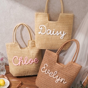 May include: Three woven straw tote bags with two handles each. The bags are light tan and brown, featuring personalized names in raised script lettering. One bag displays the name Daisy in white, another shows Chloe in pink, and the third shows Evelyn in white.