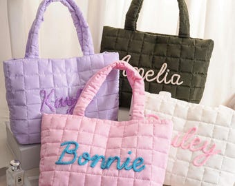 May include: Four quilted tote bags in various colors: lavender, olive green, pink, and white. Each bag features a different name embroidered in a contrasting color: Kaylee, Amelia, Bonnie, and Riley. The bags have matching padded handles.