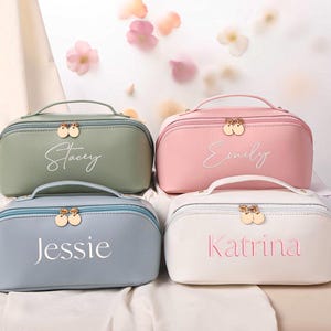 May include: Four personalized cosmetic bags in pastel colors: sage green, pink, blue, and white. Each bag has a handle, a gold zipper, and a name embroidered on the front. The names are "Stacey", "Emily", "Jessie", and "Katrina".