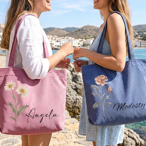 Puede incluir: Dos bolsos tote de pana, uno rosa con dise&ntilde;o floral de margaritas y el nombre "Angela", el otro azul con dise&ntilde;o floral de rosas y el nombre "Modesty". Ambos bolsos tienen asas largas.