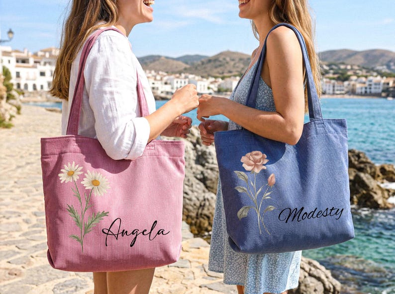 Puede incluir: Dos bolsos tote de pana, uno rosa con dise&ntilde;o floral de margaritas y el nombre "Angela", el otro azul con dise&ntilde;o floral de rosas y el nombre "Modesty". Ambos bolsos tienen asas largas.