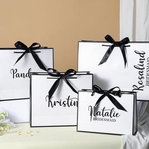 May include: Four white gift bags with black ribbon bows and black text. The bags are labelled "Pandora", "Kristin", "Natalie Bridesmaid", and "Rosalind Bridesmaid".