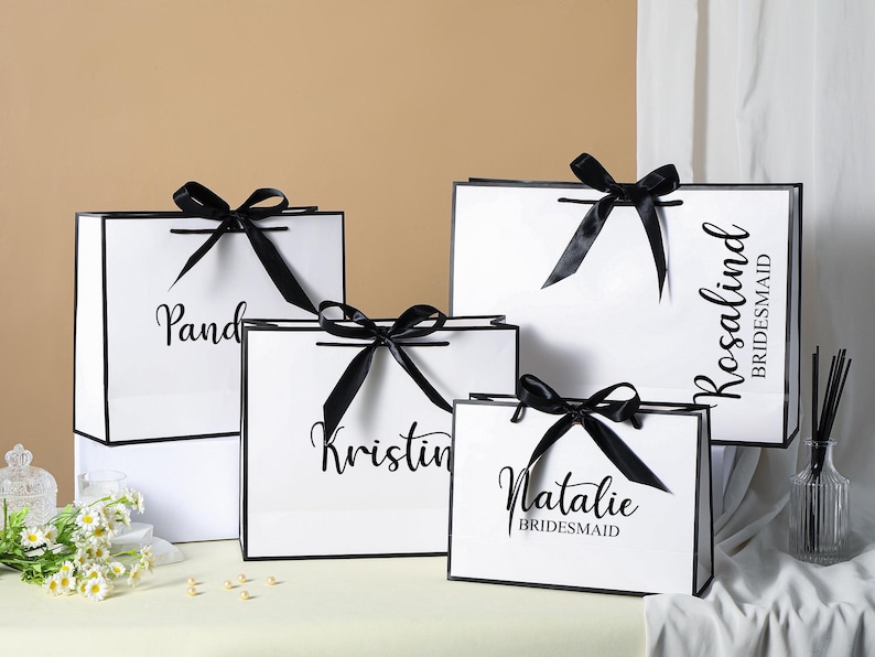 May include: Four white gift bags with black ribbon bows and black text. The bags are labelled "Pandora", "Kristin", "Natalie Bridesmaid", and "Rosalind Bridesmaid".