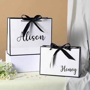 May include: Two white gift bags with black ribbon bows and black trim. The larger bag has the name "Alison" printed on it. The smaller bag has the name "Honey" printed on it.