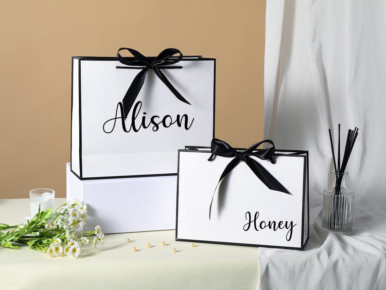 May include: Two white gift bags with black ribbon bows and black trim. The larger bag has the name "Alison" printed on it. The smaller bag has the name "Honey" printed on it.