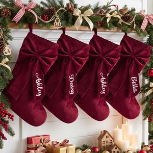 May include: Four deep red velvet Christmas stockings hanging in a row, each featuring a large matching velvet bow and a white embroidered name tag. The names Ashley, Daisy, Ashley, and Bella are written in a cursive white script on the bows.