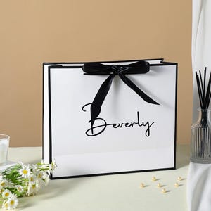 May include: White gift bag with a black ribbon and black trim. The bag has the word "Beverly" printed in black script on the front.