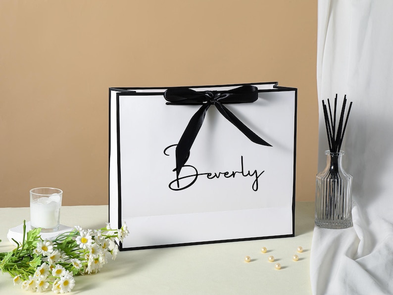 May include: White gift bag with a black ribbon and black trim. The bag has the word "Beverly" printed in black script on the front.