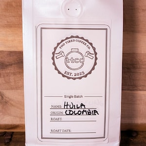 May include: A white coffee bag with a label displaying "Too Tired Coffee Co." and "EST. 2023." The label states the coffee is single-batch, named "Huila" and sourced from "Colombia." The bag is sealed with black tape, and coffee beans are scattered around.