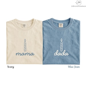 May include: Two crew neck t-shirts, one ivory and one blue jean, each featuring a graphic of a single lit birthday candle above the word mama on the ivory shirt and the word dada on the blue jean shirt, both with Comfort Colors labels.