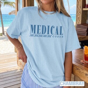 Puede incluir: Camiseta azul claro con las palabras "MEDICAL CODER" en azul oscuro. El texto está sobre un patrón de cuadros. La camiseta es de un material suave y está etiquetada como "CHAMBRAY" en la parte inferior. La camiseta se usa con pantalones a rayas azules y blancas.
