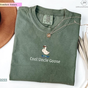 May include: A moss green crew neck t-shirt featuring an embroidered graphic of a white goose wearing sunglasses while riding a blue skateboard. Below the graphic, the text Cool Uncle Goose is printed in white. The shirt has a Comfort Colors label.