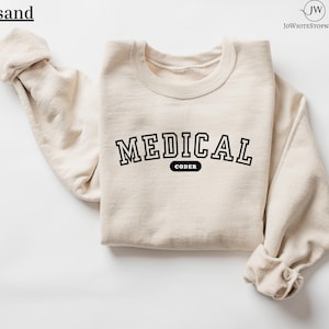 May include: A sand-colored sweatshirt with the word "MEDICAL" in bold, black, collegiate-style lettering. Below the word "MEDICAL" is the word "CODER" in a smaller, black, rectangular box. The sweatshirt is folded neatly.