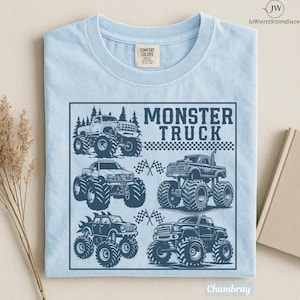 May include: Light blue t-shirt with a graphic of several monster trucks in a square frame. The words "MONSTER TRUCK" are printed in a bold font above the trucks. The shirt is made of a soft, comfortable material.