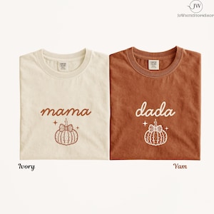 Könnte beinhalten: Zwei T-Shirts mit Rundhalsausschnitt, eines in Elfenbein und eines in Yamswurzel-Farbe, jeweils mit einem Grafikdruck. Das elfenbeinfarbene Shirt zeigt das Wort mama und das yamswurzelfarbene Shirt das Wort dada über einer Kürbisgrafik.