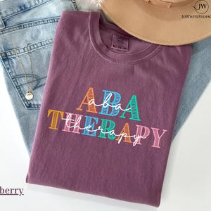 May include: A berry-coloured t-shirt with the words "ABA THERAPY" in colourful block letters and script. The shirt is folded on top of a pair of blue jeans, with a gold necklace and a tan hat visible in the background.